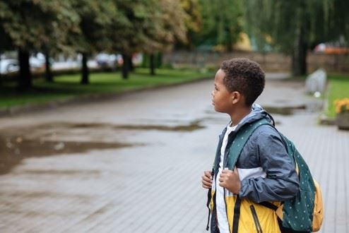 A young boy wearing a backpack stands on a wet city sidewalk.